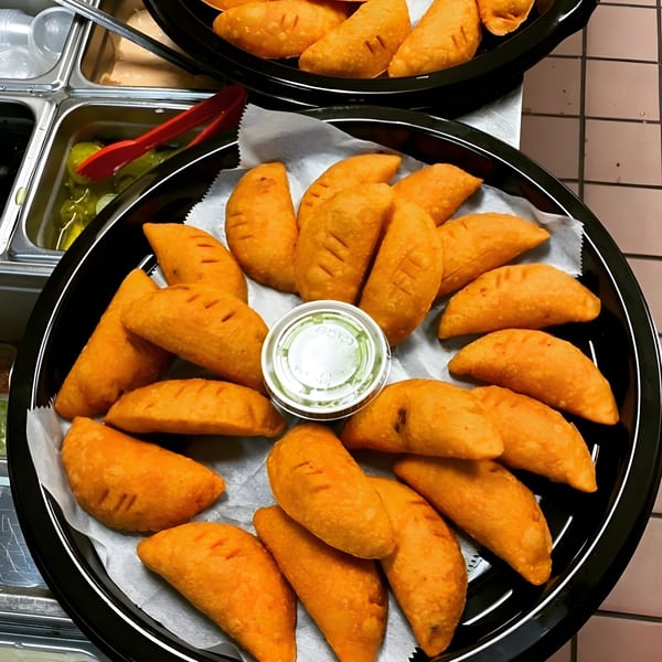 Two black trays filled with golden-brown Latin American empanadas, with a small cup of green sauce.